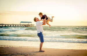 Non-surgical back pain relief therapy at Campbell Chiropractic young happy father holding up in his arms little son putting him up at the beach in barefoot standing in front of sea waves wet sand having fun with the kid in Summer sunset coast