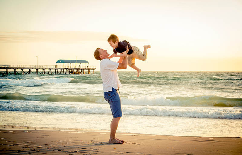young happy father holding up in his arms little son putting him Non-surgical back pain relief therapy at Campbell Chiropractic young happy father holding up in his arms little son putting him up at the beach in barefoot standing in front of sea waves wet sand having fun with the kid in Summer sunset coast