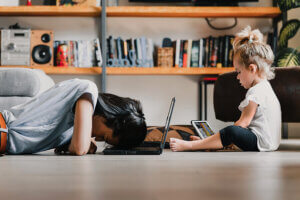 Young woman, presumably a mother, is lying on the floor with her head on a laptop, appearing exhausted, while her daughter is sitting next to her playing on a tablet