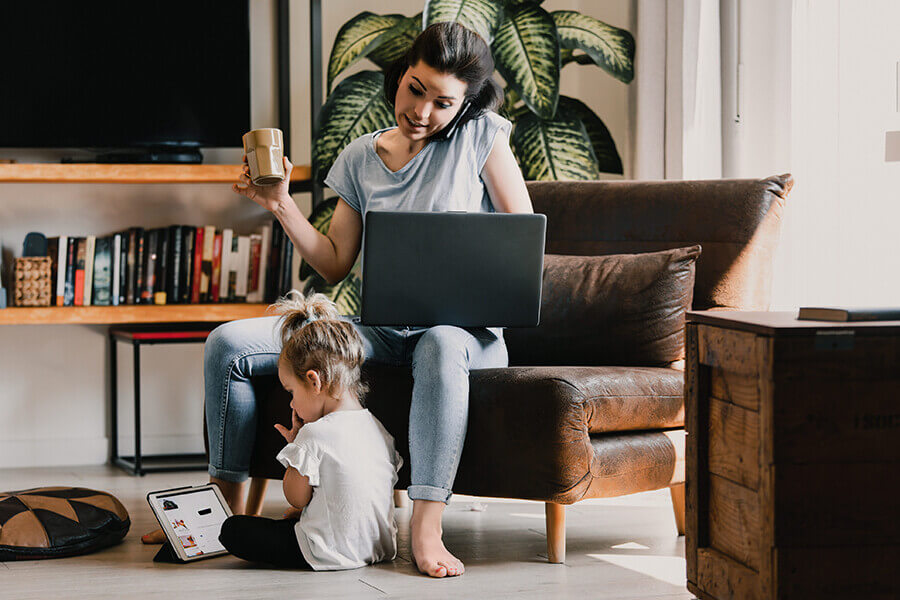Busy mother multitasks at home, balancing work and childcare, working on a laptop while her daughter plays nearby
