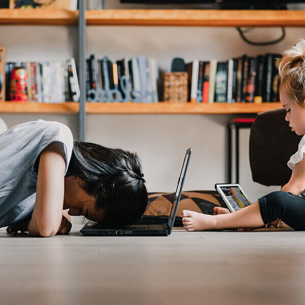 Young woman, presumably a mother, is lying on the floor with her head on a laptop, appearing exhausted, while her daughter is sitting next to her playing on a tablet